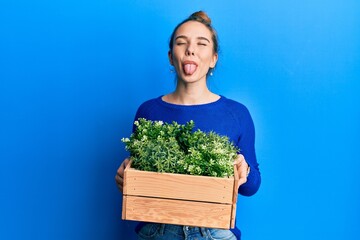 Young blonde woman holding wooden plant pot sticking tongue out happy with funny expression.
