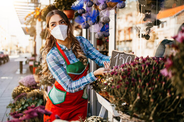 Young adult woman working in city street flower shop or florist. She is wearing protective face mask during Covid-19 virus pandemic. Small business concept.