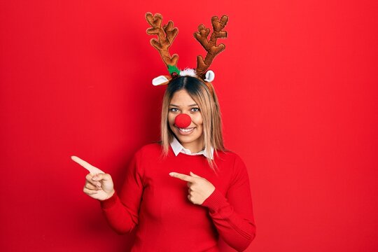Beautiful hispanic woman wearing deer christmas hat and red nose smiling and looking at the camera pointing with two hands and fingers to the side.