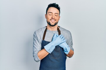 Young hispanic man wearing apron and gloves smiling with hands on chest with closed eyes and grateful gesture on face. health concept.