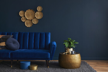 Elegant modern living room interior design with glamour blue velvet sofa, pouf, golden metal side table, plants and modern home accessories. Dark blue wall. Template. Copy space..