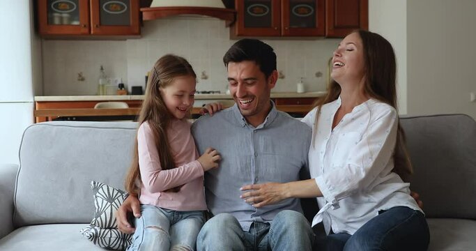 Little cute daughter tickle loving young stepfather seated together with smiling cheerful mother on sofa, enjoy playtime and communication on weekend at home. Family playtime, Happy Father Day concept