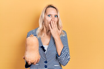 Beautiful caucasian blonde woman holding healthy fresh pumpkin covering mouth with hand, shocked...