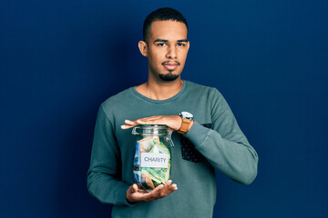 Young african american man holding charity jar with south africa rand banknotes relaxed with serious expression on face. simple and natural looking at the camera.