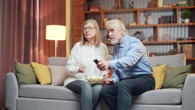 grey-haired older couple at home choose a television show to watch and switch channels with a TV remote control. elderly mature family husband and wife cannot choose a show. watching news or film