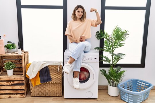 Young hispanic woman drinking coffee waiting for washing machine at laundry room strong person showing arm muscle, confident and proud of power