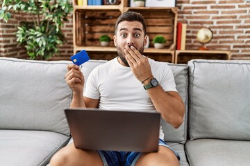 Handsome hispanic man doing online shopping holding credit card covering mouth with hand, shocked...