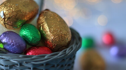 Plan rapproché sur un panier remplit d’œufs de pâque avec en arrière plan des chocolats et un fond flou lumineux.