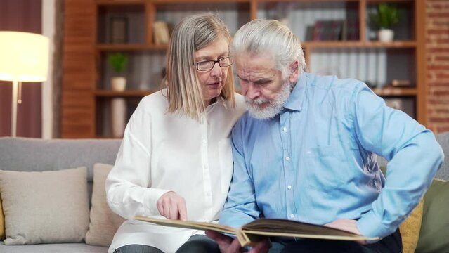 A lovely senior couple watching Memoirs photo album on sofa at home. Elderly family grandparents sitting together looking and discussing pleasant moments turnes pages with photographs, nostalgia