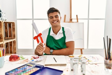 Young hispanic man at art studio holding degree smiling positive doing ok sign with hand and...