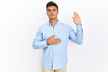 Young hispanic man wearing business shirt standing over isolated background swearing with hand on...