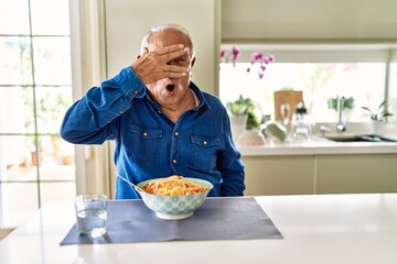 Fototapeta na wymiar Senior man with grey hair eating pasta spaghetti at home peeking in shock covering face and eyes with hand, looking through fingers with embarrassed expression.