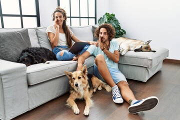 Young hispanic couple with dogs relaxing at home hand on mouth telling secret rumor, whispering malicious talk conversation