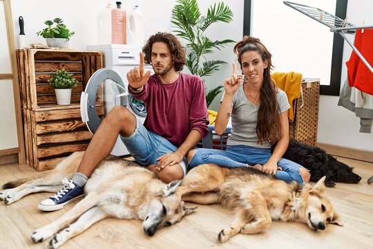 Young hispanic couple doing laundry with dogs pointing with finger up and angry expression, showing no gesture
