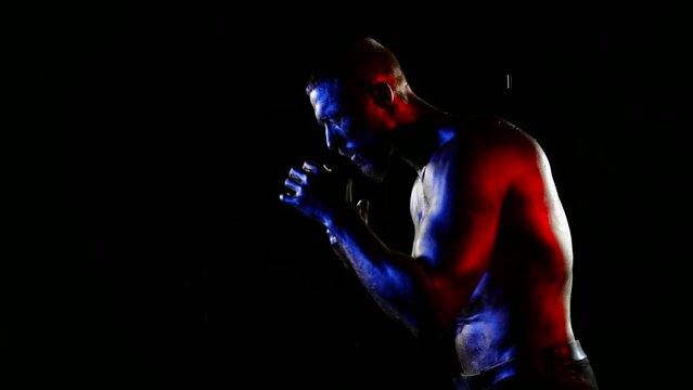 a muscular man with a bare torso and golden metallic skin fights with a shadow under water drops on a dark background. side view. the average plan. white, red and blue light