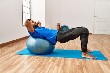 Handsome hispanic man doing exercise and stretching on yoga mat, practicing flexibility with pilates ball at the gym