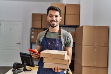 Young hispanic man business worker using smartphone holding packages at storehouse