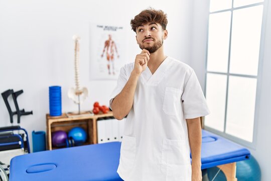 Young arab man working at pain recovery clinic with hand on chin thinking about question, pensive expression. smiling with thoughtful face. doubt concept.