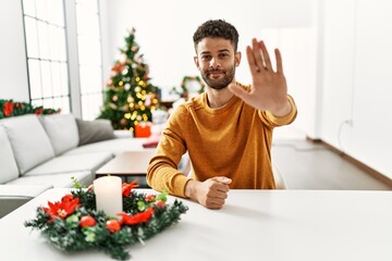 Arab young man sitting on the table by christmas tree doing stop sing with palm of the hand. warning expression with negative and serious gesture on the face.