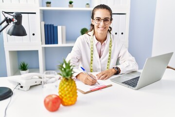 Young hispanic woman wearing nutritionist uniform writing on clipboard at clinic