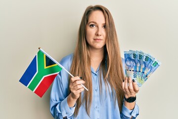 Young blonde woman holding south african flag and rands relaxed with serious expression on face. simple and natural looking at the camera.