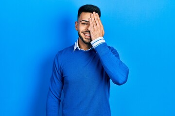 Young hispanic man with beard wearing casual blue sweater covering one eye with hand, confident smile on face and surprise emotion.