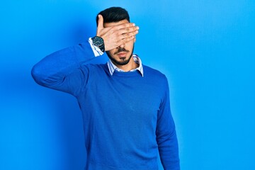 Fototapeta premium Young hispanic man with beard wearing casual blue sweater covering eyes with hand, looking serious and sad. sightless, hiding and rejection concept