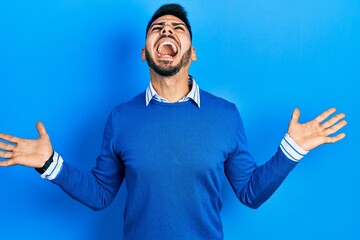 Young hispanic man with beard wearing casual blue sweater crazy and mad shouting and yelling with...