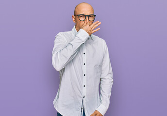 Bald man with beard wearing business shirt and glasses smelling something stinky and disgusting,...
