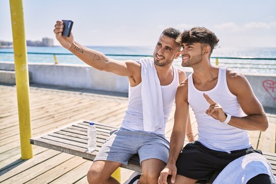 Two hispanic men sporty couple make selfie by the smartphone sitting on bench at seaside