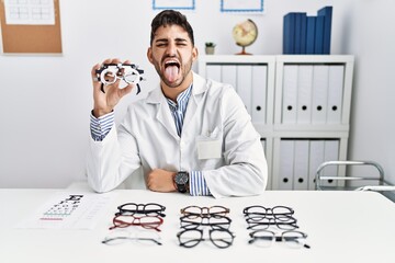 Young optician man holding optometry glasses sticking tongue out happy with funny expression. emotion concept.