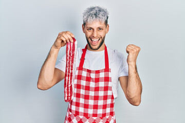 Young hispanic man with modern dyed hair wearing apron holding raw beef steak screaming proud, celebrating victory and success very excited with raised arms