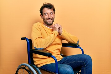 Handsome man with beard sitting on wheelchair laughing nervous and excited with hands on chin looking to the side