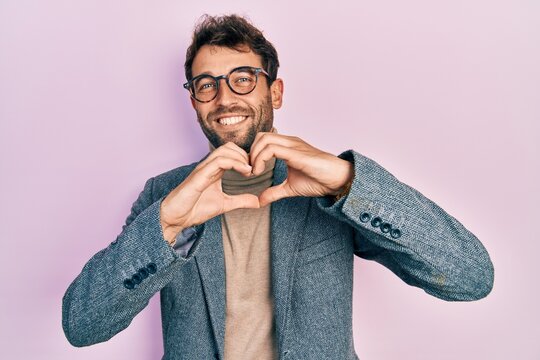 Handsome man with beard wearing business jacket and glasses smiling in love doing heart symbol shape with hands. romantic concept.