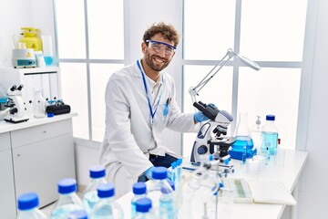 Fototapeta premium Young hispanic man wearing scientist uniform using microscope at laboratory