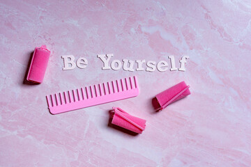 feminine Be Yourself concept with text, foam curlers and a comb on a pink marbled background