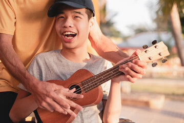 Happy face of young man with disability holding ukulele and singing, playing with music therapy on the outdoor nature background,Vacation hobby activity with family activity and mental health concept.