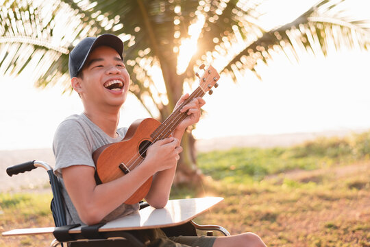 Happy Face Of Young Man With Disability Holding Ukulele And Singing, Playing With Music Therapy On The Outdoor Nature Background,Vacation Hobby Activity With Family Activity And Mental Health Concept.