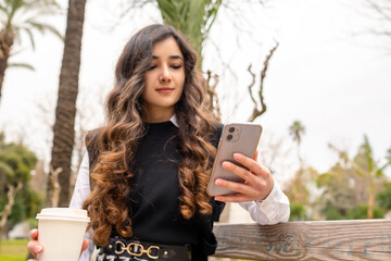 Woman with wavy hair looking at her phone, she is holding a coffee cup and reading a text on the scree, she hangs out alone in an open park, selective focus on phone