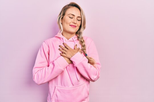 Beautiful young blonde woman wearing pink sweatshirt smiling with hands on chest, eyes closed with grateful gesture on face. health concept.