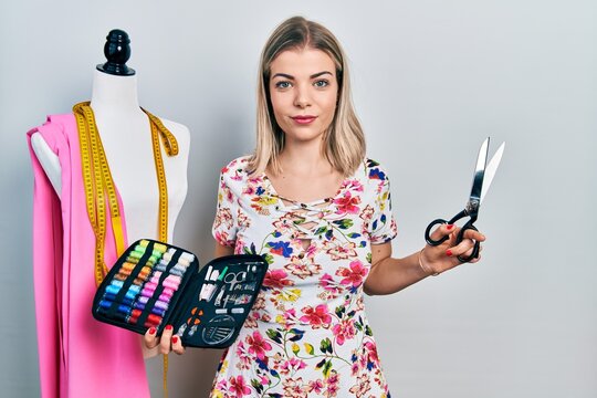 Beautiful caucasian woman dressmaker designer holding scissors and sewing kit relaxed with serious expression on face. simple and natural looking at the camera.