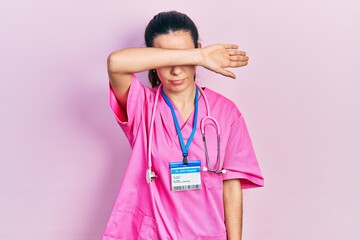 Young brunette woman wearing doctor uniform and stethoscope covering eyes with arm, looking serious and sad. sightless, hiding and rejection concept