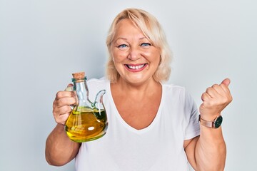 Middle age blonde woman holding olive oil can screaming proud, celebrating victory and success very excited with raised arm