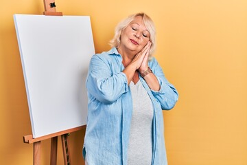 Middle age blonde woman standing by painter easel stand sleeping tired dreaming and posing with...