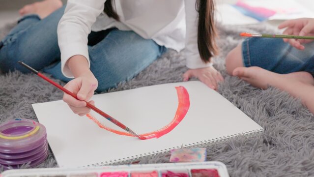 Children's combined creation in the living room as two girls paint with watercolor paints and brushes on a huge drawing paper on the floor.
