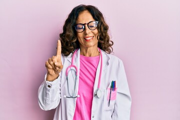 Middle age hispanic woman wearing doctor uniform and glasses showing and pointing up with finger number one while smiling confident and happy.