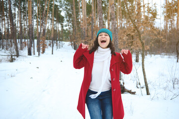 Cheerful beautiful mixed race woman in bright red coat spinning around herself feeling happy, enjoying freedom while relaxing in a snowy forest with sunbeams falling through pine trees