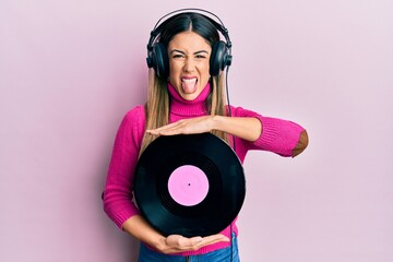 Young hispanic woman listening to music using headphones holding vinyl disc sticking tongue out...