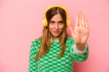 Young caucasian woman listening to music isolated on pink background standing with outstretched hand showing stop sign, preventing you.