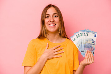 Young caucasian woman holding banknotes isolated on pink background laughs out loudly keeping hand on chest.